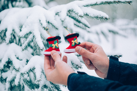 Close up man hand holding red Christmas toy Santa boots in front of snow covered fir Christmas tree, outdoors.の写真素材