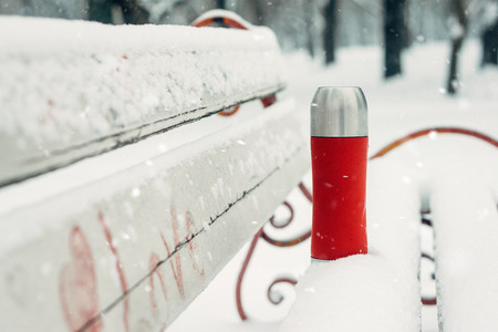 Winter Activities outdoor. Winter holidays, hot drinks concept. Red flask and cup on snowy bench in winter park.の写真素材