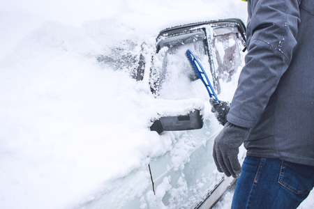 Man cleaning car from snow and ice with brush and scraper tool during snowfall. Winter emergency. Weather-related vehicle emergencies. Automobile covered with snowの写真素材