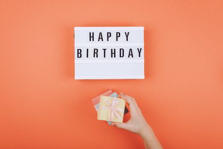 Happy birthday gift flat lay background. Children hands holding gift box with ribbon bow and light box with text Happy birthday on coral background. の写真素材
