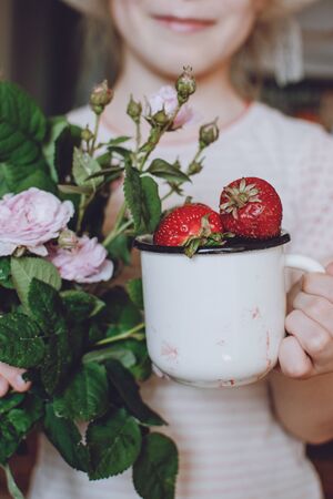Girl child holding fresh red organic strawberries in white enameled cup and bouquet of rose. Fresh organic Strawberries in children hands. Summer weekend garden background.の写真素材