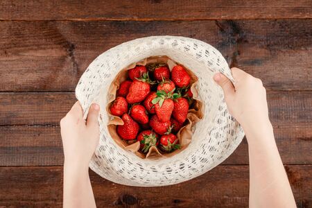 Child hand holding Fresh organic strawberries in round straw hat on brown rustic wooden background. Red berries on table flat lay.の写真素材