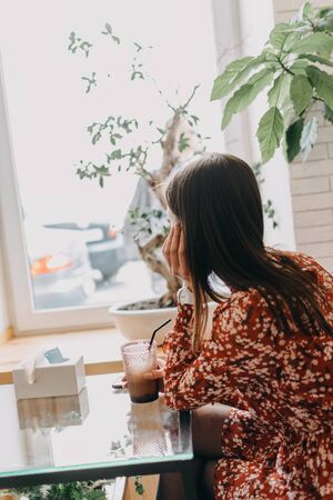 Back view of young brunette woman sitting in cafe alone drinking coffee and looking out the windowの写真素材