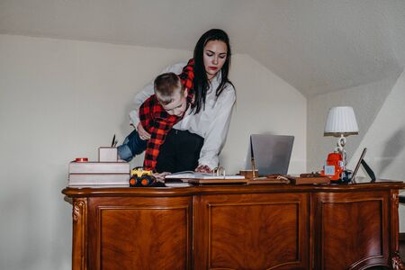 How to work from home with kids. Home office, social distancing, quarantine, isolation and parenting concept. Authentic photos of businesswoman and her son in home office work and play togetherの写真素材
