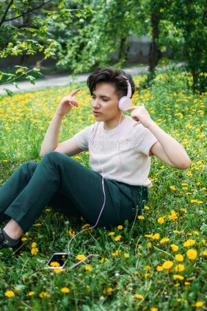 Young beautiful woman sitting on the lawn green grass with dandelions and listen to music in headphones. Young gen z girl listening to music on headphones in a summer park.の写真素材