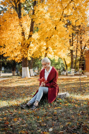 Beautiful blonde Woman with Autumn Leaves on Fall Nature Background. Alone brooding woman in red coat holding autumn leafs in the nature.の写真素材