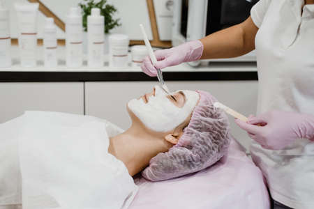 Cosmetologist applying mask on client's face in spa salon. Young woman getting facial care by beautician at spa salon Acne Treatment, face peeling mask, spa beauty treatment, skincare.の写真素材