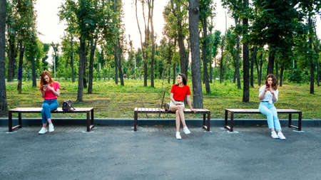 Social distancing, new normal concept. Three woman girlfriends with cell phone sitting on bench in park in social distancing. Social distance to avoid the spread of coronavirusの写真素材