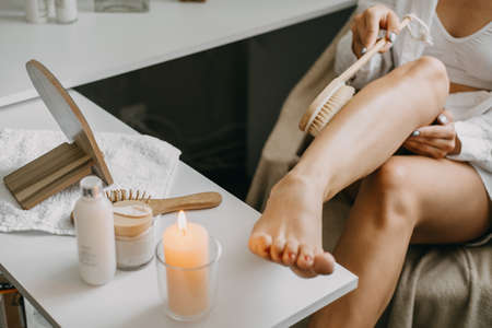 Dry brushing body brush for exfoliating dry skin, lymphatic drainage and cellulite treatment. Woman brushing skin leg with dry wooden brush to prevent cellulite and body problem in bedroom at homeの写真素材