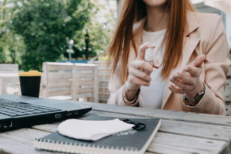 Working safely in new normal. How to make a COVID secure workplace. Redhead Business woman hair working in street cafe fresh air office with laptop, face mask and sanitizerの写真素材
