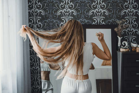How take care of long hair. Back view of young natural blonde woman in white home clothes combing her hair and looking into the mirror in bedroomの写真素材