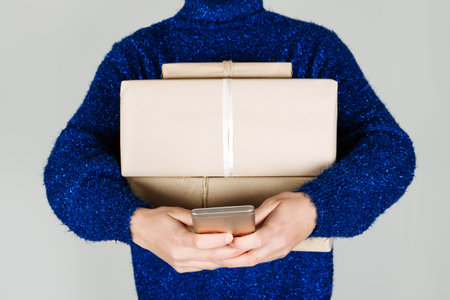 Winter Christmas online shopping banner with female hands with many craft gift boxes and cell phone. Winter Christmas sale background with woman in blue sweater holding lot of boxes and phone in handsの写真素材