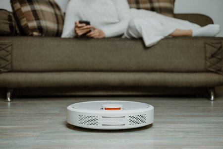 Robotic Vacuums, Robot Mops. Smart home. Robotic Vacuum Cleaner while Woman Relaxing on sofa. Automatic robotic vacuum cleaner on laminate wood floor, smart cleaning technologyの写真素材