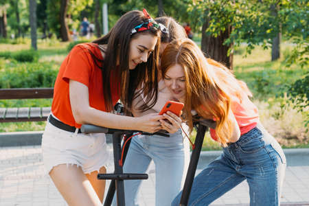 Three young girl friends on the electro scooters taking picture of themselves on cell phone at summer sunny day. Outdoor portrait of three friends girl riding electric kick scooter in the parkの写真素材