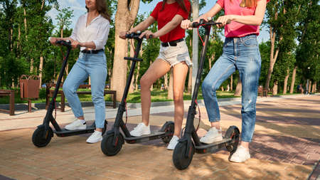 Three young girl friends on the electro scooters having fun in city street at summer sunny day. Outdoor portrait of three friends girl riding electric kick scooter in the parkの写真素材