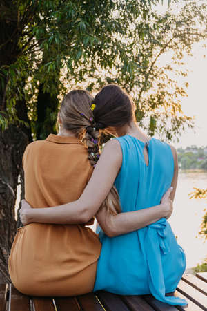 Female friendship femininity, diverse, support, partnership, girlfriend concept. Two girls blonde, brunette, with braided plait sitting and hugging in the nature sunset backgroundの写真素材