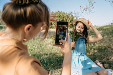 Back view of Young woman holding cell phone and making photo of her girl friend. Two girlfriends take pictures of each other on Smartphone camera on nature background. Summer holidays and technologyの写真素材