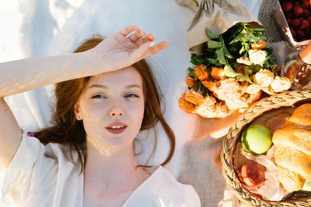 Top view candid portrait of young red-haired woman lying on a picnic blanket near flowers and basket...