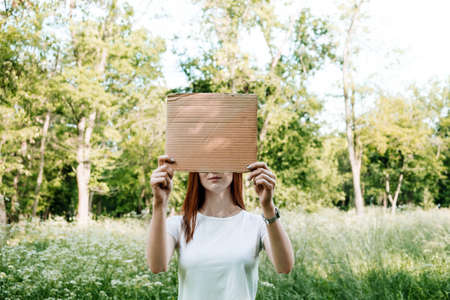 Outdoor mockup Young redhead woman covers her face with cardboard poster. Faceless mock up portrait of woman holding cardboard sign on nature background.の写真素材