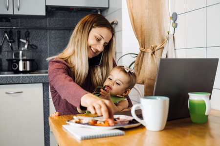 Working mother work remotely. Remote Work from home. Young mother with toddler baby girl working at home using laptop on kitchen backgroundの写真素材