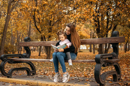 Young female woman babysitter and toddler baby girl read book in autumn park. Happy family mom and toddler outdoors in fall parkの写真素材