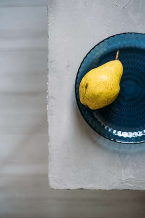 Close up one ripe yellow pear in blue glass plate on grey background. Modern minimal still life with one yellow pear. Copy spaceの写真素材