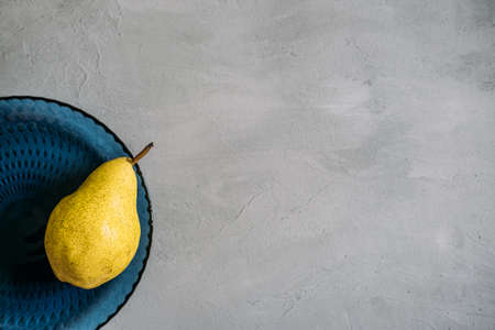Close up one ripe yellow pear in blue glass plate on grey background. Modern minimal still life with one yellow pear. Copy spaceの写真素材