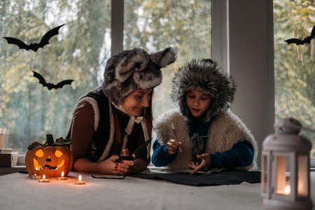 Mom and son in Halloween costumes play together and make paper bat Halloween decorations on pumpkins lamp at home backgound. Halloween family at home, Halloween parties and decorationsの写真素材