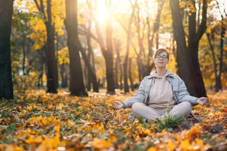Caucasian middle aged woman meditating in lotus pose at autumn park with sunlight. Yoga at fall park. Woman in glasses relaxing on yellow leaves. Meditation, Mental health, self care, mindfulnessの写真素材