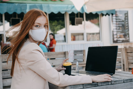 Redhead Business woman wearing face mask for virus protection working on the laptop outdoors. Young...