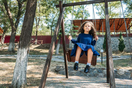 Happy little girl in blue dress on swing in summer park. Cute girl in the park swinging on a swing. Summer active leisure and activity outdoorsの写真素材