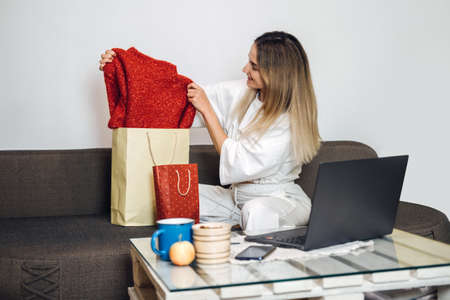 Black friday, online sale, internet shopping, ecommerce. Caucasian woman pulls new clothes out of shopping bag on laptop background at home. Woman enjoy weekend shopping home indoorsの写真素材