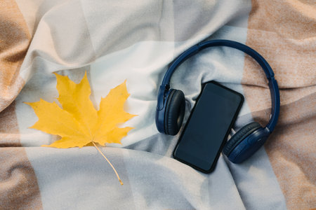 Smartphone mockup, headphones, yellow maple leaf on blanket in autumn park background. Audio technology apps, meditation application, fall mood music podcasts and booksの写真素材