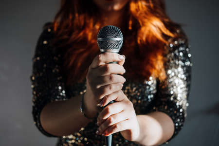 Faceless portrait of redhead woman in sparkly evening dress holding microphone on dark night background. Unrecognizable female singer with modern microphone in handsの写真素材