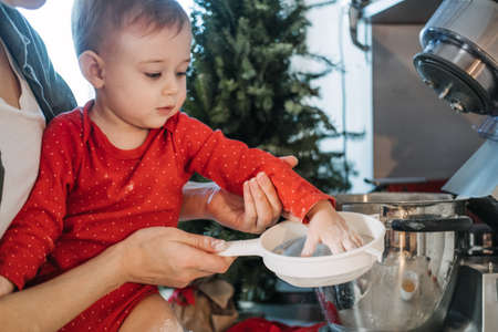 Cozy Christmas at home. Family preparation holiday dessert. Mother and baby toddler daughter play with flour, make dough and cooking gingerbread together on kitchen at home.の写真素材