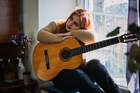 Unaltered candid portrait of young red haired woman in sweater playing acoustic guitar sitting by...