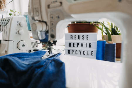Reuse, repair, upcycle text on light board on sewing machines background. Stack of old jeans, Denim clothes, scissors, thread and sewing tools in sewing studio. Denim Upcycling Ideas, Using Old Jeansの写真素材