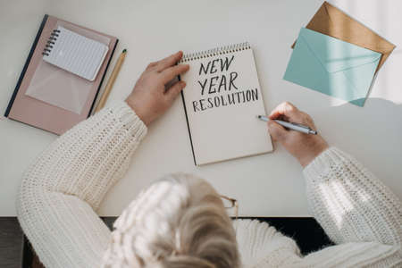 Healthy New Years resolutions for older adults. Senior mature old woman in white sweater writing handwritten text New Years resolutions in open notepad on the table. Planning and setting goalsの写真素材