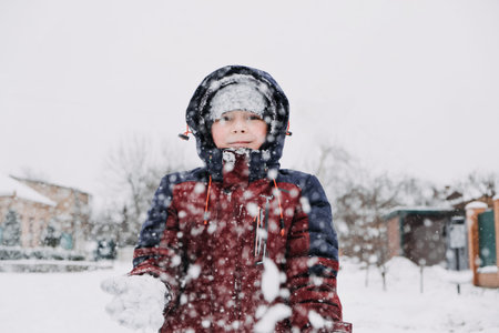 Close up outdoor winter portrait of boy playing snowballs. Authentic, real, candid portrait of cute boy in winter time. Little kid in winter clothes walking under the snowの写真素材