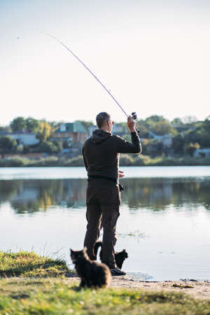 Fisherman with spinning rod and cat near him on nature background. Angler man with fishing spinning or casting rod by the river. Fisherman with rod, spinning reel on the river bankの写真素材