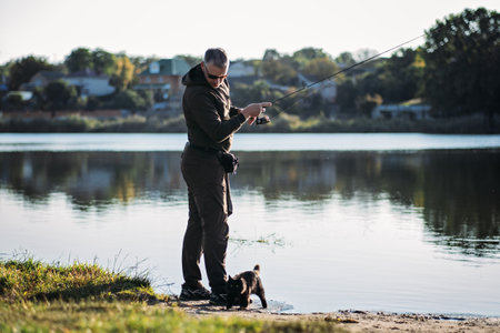 Fisherman with spinning rod and cat near him on nature background. Angler man with fishing spinning or casting rod by the river. Fisherman with rod, spinning reel on the river bankの写真素材
