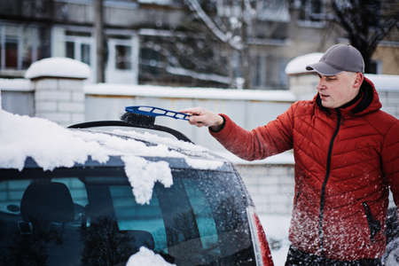 Man cleaning car from snow and ice with brush and scraper tool during snowfall. Winter emergency. Weather-related vehicle emergencies. Automobile covered with snowの写真素材