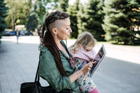 Freelance Parents. Busy working mom with baby toddler daughter on hands walk and looking on tablet in street. Multitasking Freelancer mom making notes while holding her baby outddors.の写真素材