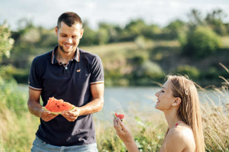 Young couple in love on summer picnic with watermelon. Loving couple sitting by the river, talking, smiling, laughing, eating watermelonの写真素材