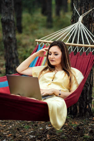 Young happy successful woman freelancer with laptop relaxing in the hammock on summer Pine forest on sunny day. Female entrepreneur working with laptop in hammock and camping in nature.の写真素材