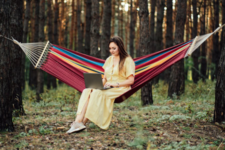 Young happy successful woman freelancer with laptop relaxing in the hammock on summer Pine forest on...