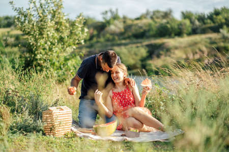 Young couple in love on summer picnic with watermelon. Loving couple sitting by the river, talking, smiling, laughing, eating watermelonの写真素材
