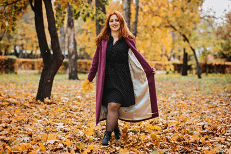 Outdoor autumn portrait of happy smiling plus size red hair woman in coat walking in fall park.の写真素材