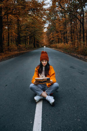 Confidence woman with book sitting on the road with autumn tree around. New life, path choice, ambitionの写真素材