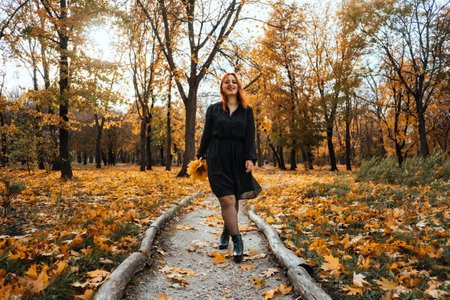 Outdoor autumn portrait of happy smiling plus size red hair woman in dress walking in fall park. Candid portrait of happy curvy womanの写真素材